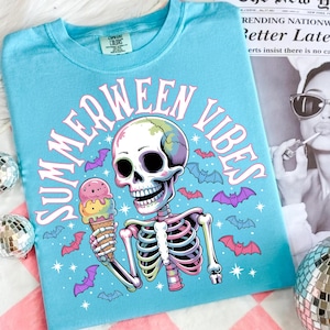 May include: A light blue t-shirt with the words "SUMMERWEEN VIBES" in pink. The graphic features a skeleton holding an ice cream cone, surrounded by bats. The shirt is a Comfort Colors brand.