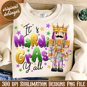 May include: Cream-colored sweatshirt with the text "It's Mardi Gras y'all" in purple, yellow, and green. A nutcracker with a gold crown and a mustache is featured. The design includes star accents.