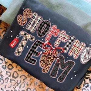 Peut inclure: Sweat-shirt gris fonc&eacute; avec l'inscription "SHOPPING TEAM" dans diff&eacute;rents motifs, dont du tartan et un imprim&eacute; l&eacute;opard, avec un n&oelig;ud rouge. Une petite &eacute;tiquette rouge avec le mot "sale" est attach&eacute;e. Le sweat-shirt est pli&eacute;.