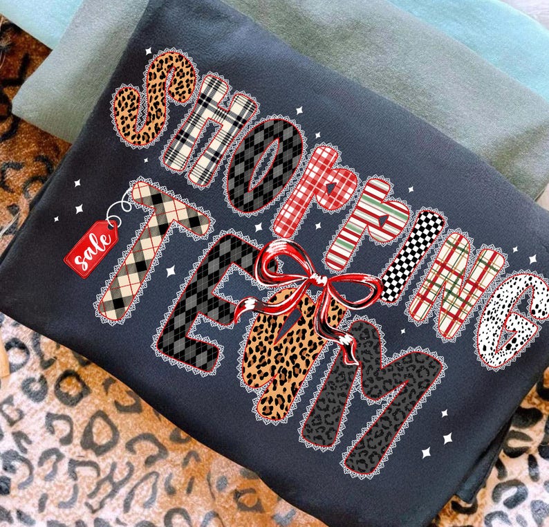 Peut inclure: Sweat-shirt gris fonc&eacute; avec l'inscription "SHOPPING TEAM" dans diff&eacute;rents motifs, dont du tartan et un imprim&eacute; l&eacute;opard, avec un n&oelig;ud rouge. Une petite &eacute;tiquette rouge avec le mot "sale" est attach&eacute;e. Le sweat-shirt est pli&eacute;.