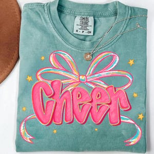 May include: A teal Comfort Colors t-shirt with a pink and rainbow bow graphic and the word "Cheer" in pink. Gold star accents surround the graphic. A silver necklace with a pendant is draped across the shirt.
