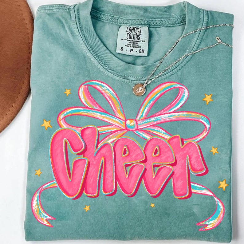 May include: A teal Comfort Colors t-shirt with a pink and rainbow bow graphic and the word "Cheer" in pink. Gold star accents surround the graphic. A silver necklace with a pendant is draped across the shirt.