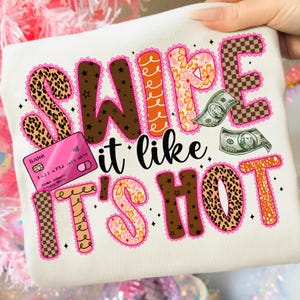 以下が含まれることがあります： クリーム色のTシャツ。 「SWIPE it like it's HOT」の文字が様々なフォントとパターンで描かれています。 ピンクのクレジットカード、お札、装飾的な要素が含まれています。