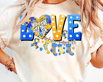 May include: A white t-shirt with the word "LOVE" in blue, yellow, and white polka dots. The "O" is a heart shape with a blue and yellow puzzle piece design and a black and white polka dot bow. There are blue and yellow puzzle pieces, a blue ribbon, and blue and yellow socks hanging from the letters.