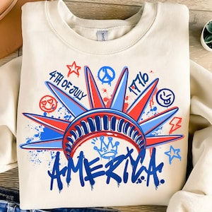 May include: Cream-colored sweatshirt with a graphic design featuring the Statue of Liberty's crown in red and blue, with the word "AMERICA" in blue graffiti-style lettering. The design includes the text "4th of July" and "1776".