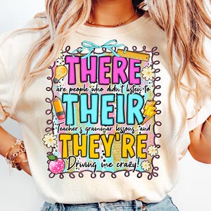 Puede incluir: Camiseta color crema con un gráfico colorido. El gráfico presenta las palabras "THERE, THEIR, THEY'RE" en letras grandes y delineadas, junto con la frase "are people who didn't listen to Teacher's grammar lessons... Driving me crazy!"