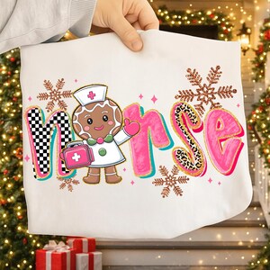 May include: White tote bag featuring the word "Nurse" in pink, with a gingerbread nurse character and snowflake accents. The "N" has a black and white checkered pattern, and the "S" has a leopard print design. A cute and festive design.