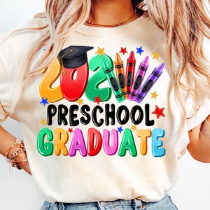 May include: A white t-shirt with a colorful graphic design that says "2023 Preschool Graduate". The design features a graduation cap, crayons, and stars.