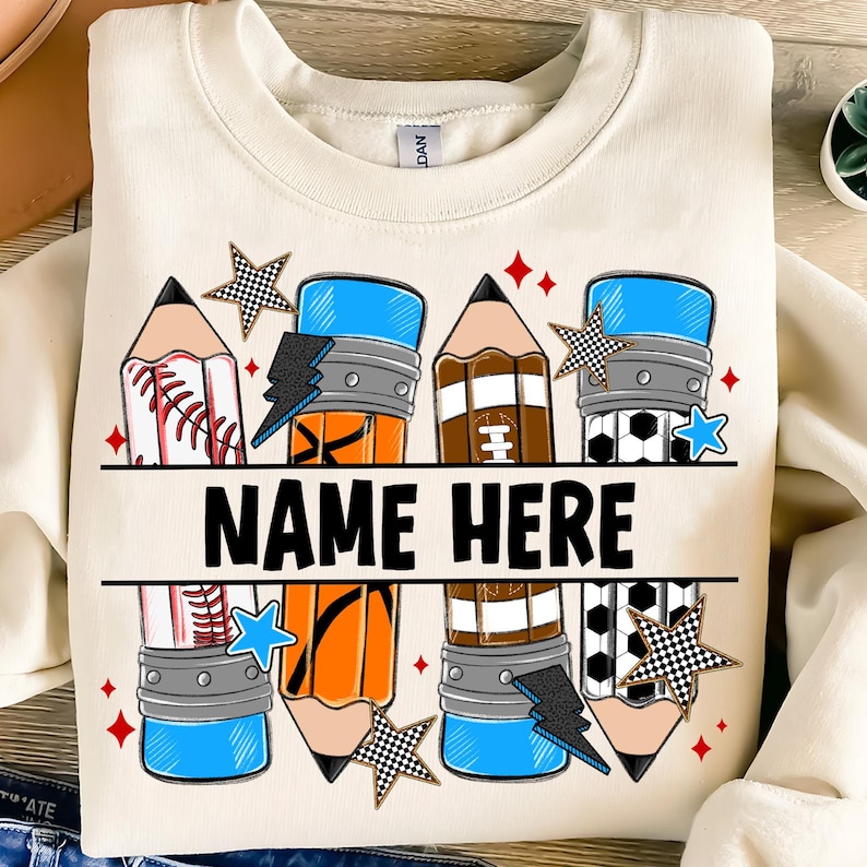 Pode incluir: Camisola creme com um design de l&aacute;pis colorido. Os l&aacute;pis apresentam temas de beisebol, basquete, futebol americano e futebol, com o texto "NAME HERE" em letras pretas em negrito. Acentuado com estrelas e raios.