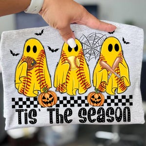 Puede incluir: Camiseta gris claro con tres figuras de fantasmas amarillos con costuras de b&eacute;isbol, cada una sosteniendo un art&iacute;culo de b&eacute;isbol. Murci&eacute;lagos negros y una telara&ntilde;a est&aacute;n encima de los fantasmas. Debajo, calabazas naranjas y el texto "Tis The Season" est&aacute;n impresos.