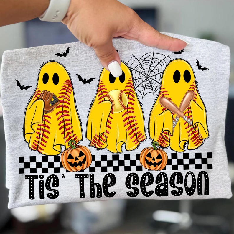Puede incluir: Camiseta gris claro con tres figuras de fantasmas amarillos con costuras de b&eacute;isbol, cada una sosteniendo un art&iacute;culo de b&eacute;isbol. Murci&eacute;lagos negros y una telara&ntilde;a est&aacute;n encima de los fantasmas. Debajo, calabazas naranjas y el texto "Tis The Season" est&aacute;n impresos.