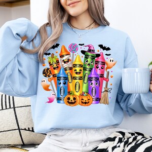 May include: Light blue sweatshirt featuring a colorful Halloween-themed crayon design. The graphic includes smiling crayons dressed as Halloween characters, pumpkins, ghosts, bats, and candy. A fun and festive design for the spooky season.