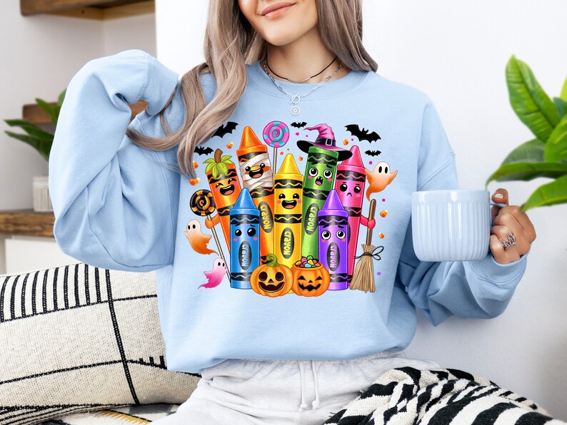 May include: Light blue sweatshirt featuring a colorful Halloween-themed crayon design. The graphic includes smiling crayons dressed as Halloween characters, pumpkins, ghosts, bats, and candy. A fun and festive design for the spooky season.