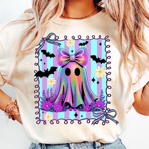 May include: A cream-colored t-shirt featuring a colorful ghost design. The ghost has a large bow and is surrounded by bats, flowers, and a striped background. The design is framed with a decorative border, perfect for Halloween.