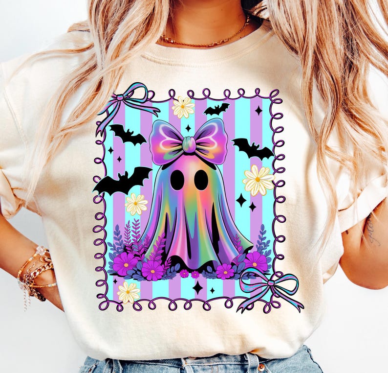 May include: A cream-colored t-shirt featuring a colorful ghost design. The ghost has a large bow and is surrounded by bats, flowers, and a striped background. The design is framed with a decorative border, perfect for Halloween.