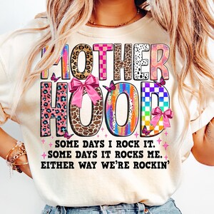 May include: A white t-shirt with a colorful graphic that says "MOTHERHOOD" with pink bows and butterflies. The text below the graphic says "SOME DAYS I ROCK IT. SOME DAYS IT ROCKS ME. EITHER WAY WE'RE ROCKIN'."