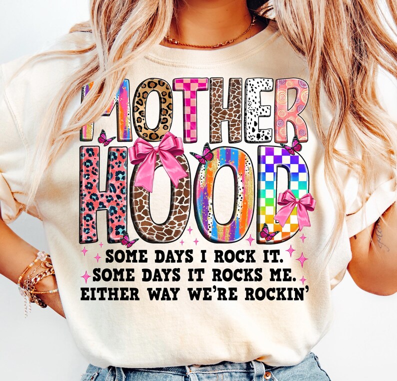 May include: A white t-shirt with a colorful graphic that says "MOTHERHOOD" with pink bows and butterflies. The text below the graphic says "SOME DAYS I ROCK IT. SOME DAYS IT ROCKS ME. EITHER WAY WE'RE ROCKIN'."