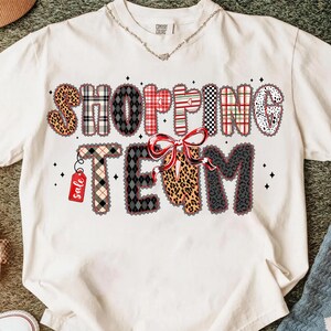 Peut inclure: T-shirt cr&egrave;me avec l'inscription "SHOPPING TEAM" dans une police d&eacute;corative. Les lettres pr&eacute;sentent divers motifs, dont un imprim&eacute; l&eacute;opard, du tartan et un n&oelig;ud. Une petite &eacute;tiquette "sale" est suspendue &agrave; la lettre "T".