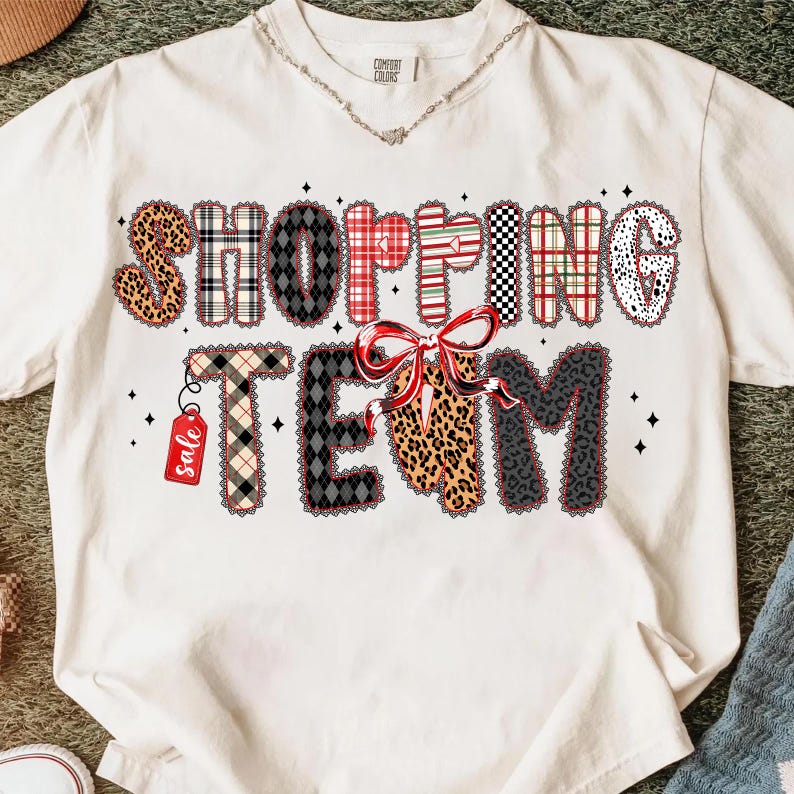 Peut inclure: T-shirt cr&egrave;me avec l'inscription "SHOPPING TEAM" dans une police d&eacute;corative. Les lettres pr&eacute;sentent divers motifs, dont un imprim&eacute; l&eacute;opard, du tartan et un n&oelig;ud. Une petite &eacute;tiquette "sale" est suspendue &agrave; la lettre "T".