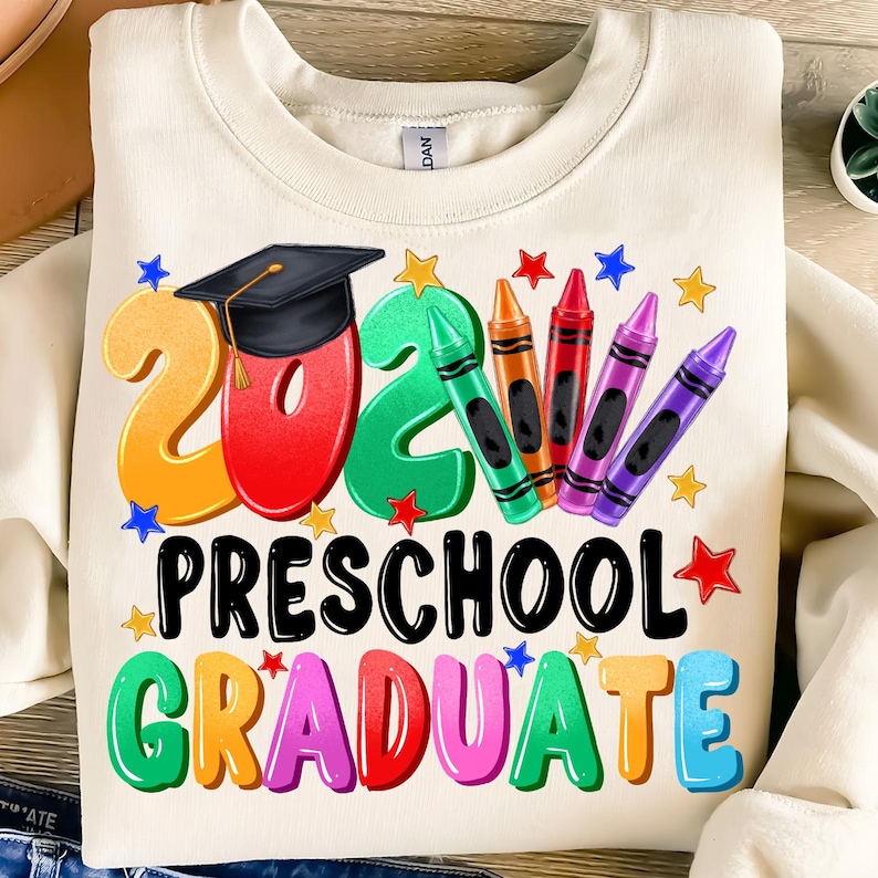 May include: A white sweatshirt with the text "2021 Preschool Graduate" in colorful letters. The number 2021 is in large, colorful letters with a graduation cap on top of the 2. There are colorful crayons and stars around the text.