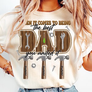 May include: Cream-colored t-shirt with a graphic design for Father's Day. The design features the word "DAD" in a wooden-style font, with tools incorporated into the letters. The text "When it comes to being the best, you nailed it" is also included.