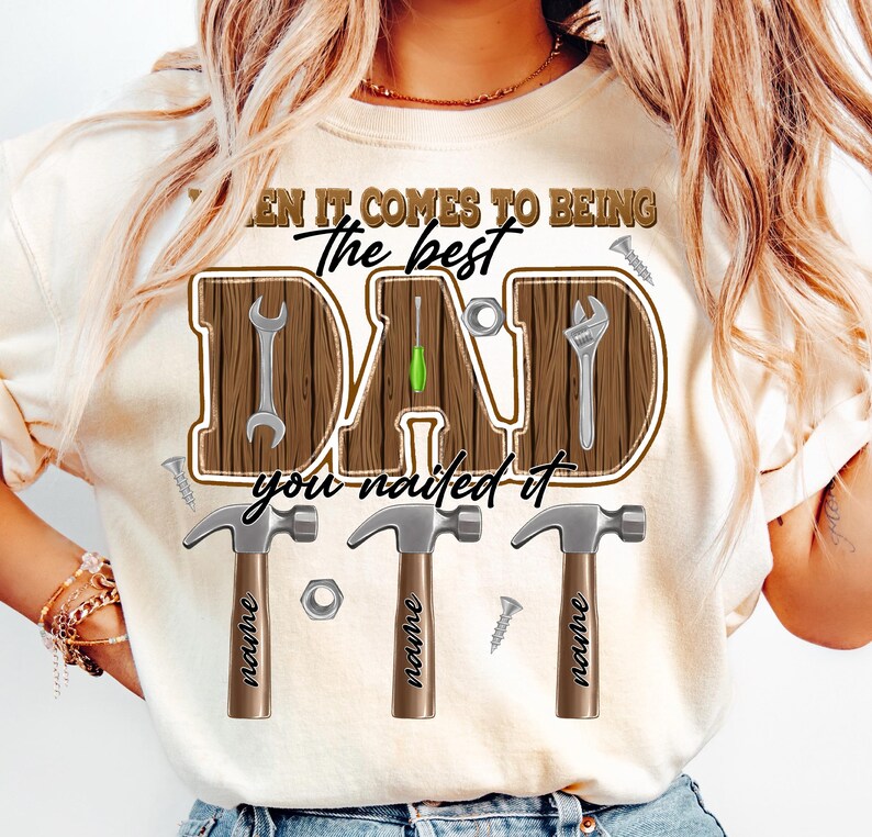 May include: Cream-colored t-shirt with a graphic design for Father's Day. The design features the word "DAD" in a wooden-style font, with tools incorporated into the letters. The text "When it comes to being the best, you nailed it" is also included.