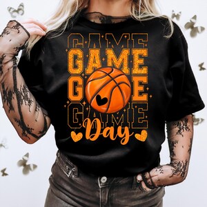 Peut inclure: T-shirt noir avec un texte en paillettes orange qui dit "Game Game Go Game Day" avec un graphique de ballon de basket orange au centre.