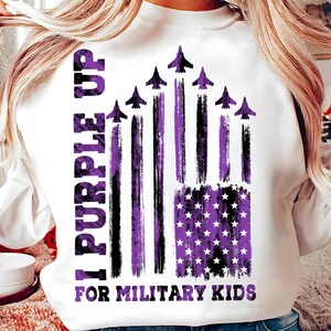 May include: White sweatshirt with a purple and black American flag design. The flag has purple stripes and stars. The text "I Purple Up For Military Kids" is printed on the sweatshirt.