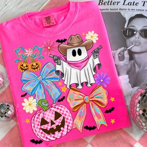 May include: Bright pink Comfort Colors t-shirt featuring a cartoon ghost cowboy with guns, a brown hat, and a pink bandana. The shirt is decorated with bows, pumpkins, flowers, bats, and stars, perfect for Halloween.