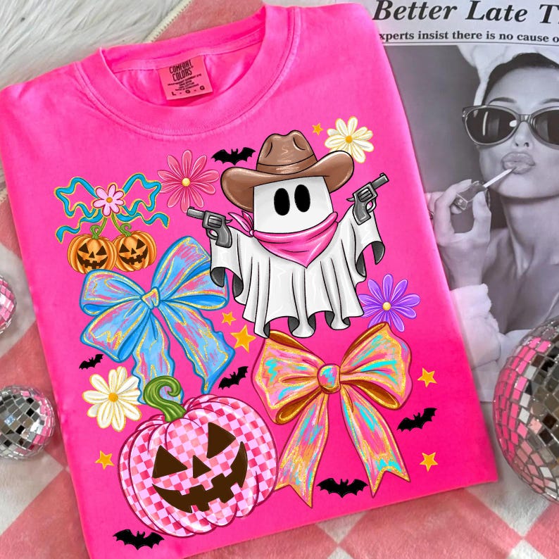 May include: Bright pink Comfort Colors t-shirt featuring a cartoon ghost cowboy with guns, a brown hat, and a pink bandana. The shirt is decorated with bows, pumpkins, flowers, bats, and stars, perfect for Halloween.