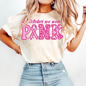 May include: A white t-shirt with the text "In October we wear PINK" in pink letters with glitter and pink ribbon, hearts, and animal print designs.