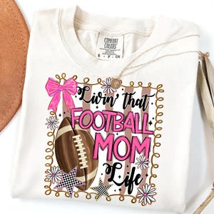 May include: Cream-colored t-shirt with a graphic that reads "Livin' that FOOTBALL MOM Life." The design includes a football, pink bow, stars, and flowers. A gold necklace is draped across the shirt.
