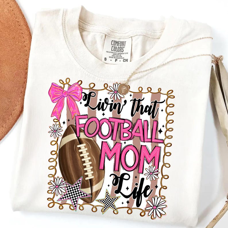 May include: Cream-colored t-shirt with a graphic that reads "Livin' that FOOTBALL MOM Life." The design includes a football, pink bow, stars, and flowers. A gold necklace is draped across the shirt.