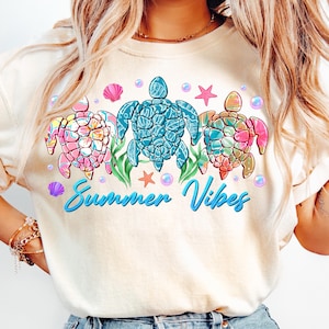 May include: A white t-shirt with a graphic of three colorful sea turtles with the text "Summer Vibes" in blue.