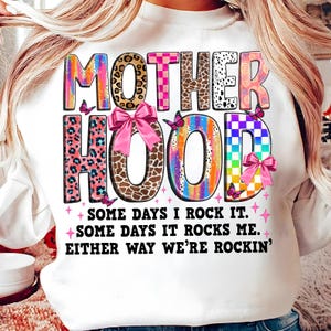 May include: White sweatshirt with a colorful graphic that says "MOTHERHOOD" with a pink bow and butterflies. The text below the graphic says "SOME DAYS I ROCK IT. SOME DAYS IT ROCKS ME. EITHER WAY WE'RE ROCKIN'."
