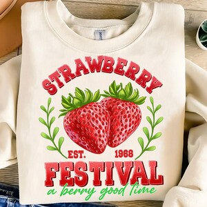 May include: A white sweatshirt with a red and green graphic design. The graphic features two strawberries, a green leafy branch, and the text "Strawberry Festival a berry good time" with the words "Est. 1968" in between.