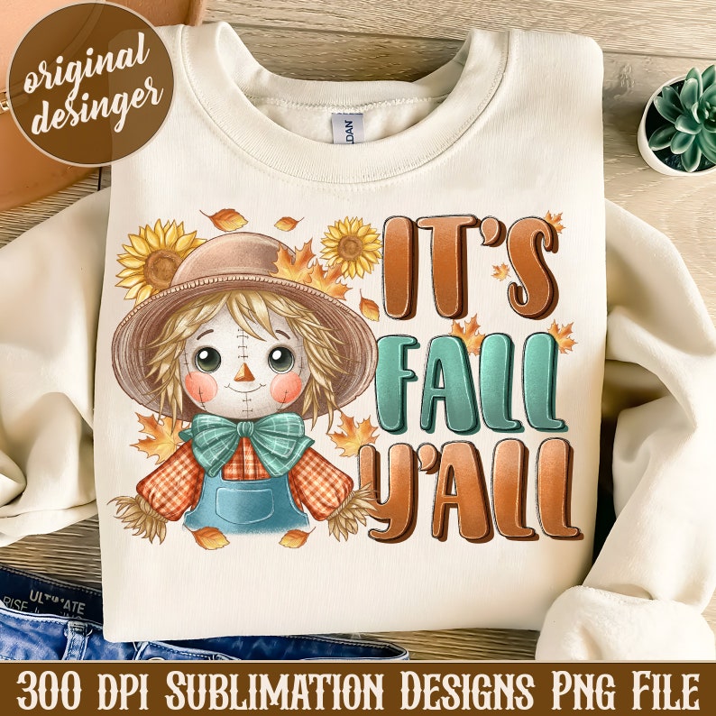 Op de afbeelding: Een wit sweatshirt met een schattig cartoon-stroopop en de tekst "It's Fall Y'all" in bruine en groene letters. De stroopop draagt ​​een bruine hoed, een rood-wit geruit overhemd en een blauwe overall. Er zijn zonnebloemen en herfstbladeren rond de stroopop.