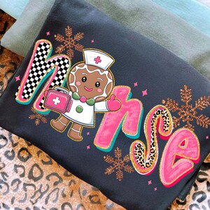 May include: Dark gray sweatshirt with the word "Nurse" in a playful font, featuring a gingerbread nurse character holding a medical bag. The letter "M" has a black and white checkered pattern, and the letter "S" has a leopard print.