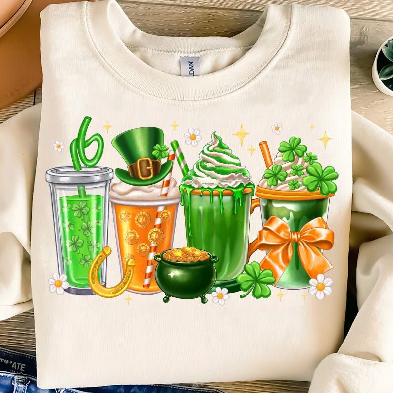 May include: A white sweatshirt with a green, orange, and white design of four different drinks with shamrocks and a gold horseshoe. The drinks include a green smoothie, a green milkshake, a green beer, and a green iced coffee.