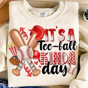 May include: A white sweatshirt with a baseball-themed design. The text "It's a Tee-ball Kinda Day" is printed in black on the front of the shirt. The design includes two crossed baseball bats, a baseball, a popcorn bucket, a baseball glove, and a baseball cap.