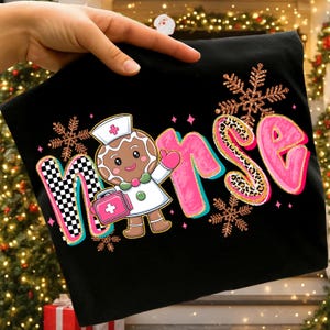 May include: Black t-shirt with a gingerbread nurse graphic. The word "Nurse" is in pink with gold glitter trim and a leopard print "S". The "M" has a black and white checkered pattern. Includes snowflakes.
