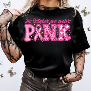 May include: Black t-shirt with pink glitter lettering that reads "In October we wear PINK". The lettering is decorated with pink hearts, a pink ribbon, and a zebra print.
