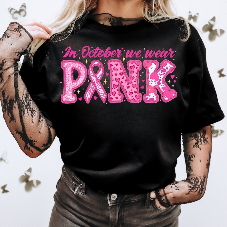 May include: Black t-shirt with pink glitter lettering that reads "In October we wear PINK". The lettering is decorated with pink hearts, a pink ribbon, and a zebra print.