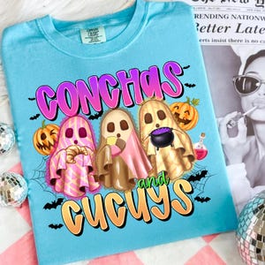 May include: A light blue t-shirt with a Halloween-themed graphic. The design features three ghost characters, pumpkins, bats, and the words "CONCHAS and CUCUYS" in a colorful font. The shirt has a crew neck and short sleeves.