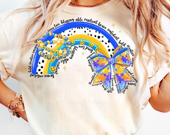 Pode incluir: Uma camiseta branca com um design de arco-íris que apresenta as palavras "happy love blessing able resilient brave confident bold creative unique" e um laço azul e amarelo com peças de quebra-cabeça. O laço tem as palavras "thoughtful accepting", "mindful", "passionate", "sincere", "joyful", "trusting", "helpful", "focused", "loving", "creative", "unique", "bold", "confident", "brave", "brilliant", "kind", "and", "love", "blessing", "able", "resilient", "brave", "confident", "bold", "creative", "unique" escritas ao redor.