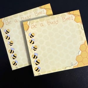 May include: Two square notepads with a yellow honeycomb pattern and cartoon bees. The text "Be a busy Bee!" is written at the top. The notepads are light yellow with a black background.