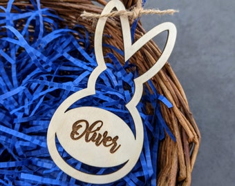 May include: A wooden bunny-shaped tag with the name "Oliver" engraved, suspended above blue shredded paper in a wicker basket. The tag is attached with twine. The image also includes the text "DIGITAL DOWNLOAD" and file type abbreviations.
