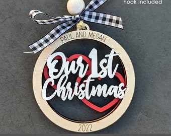 Puede incluir: Un adorno navideño de madera con una cinta a cuadros rojos y blancos. El adorno es redondo y tiene el texto "Our 1st Christmas 2022". El adorno también tiene dos corazones rojos.
