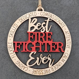 May include: A round wooden ornament with the text "Best Firefighter Ever" in red and brown lettering. The ornament is decorated with the words "Calm Under Pressure - Caring - Flexible - Skilled - Dedicated - Compassionate - Attentive".