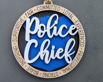 Puede incluir: Un adorno redondo de madera con una placa de policía azul en el centro. La placa tiene las palabras "Police Chief" escritas en blanco. El adorno está rodeado por un círculo de palabras que describen cualidades positivas, incluyendo "valiente", "fuerte", "profesional", "experimentado", "leal", "comprometido", "justo", "inspirador" y "solidario".